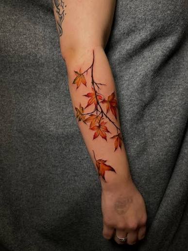 Fiery maple leaves drift along slim black branches in a vertical inner forearm tattoo.