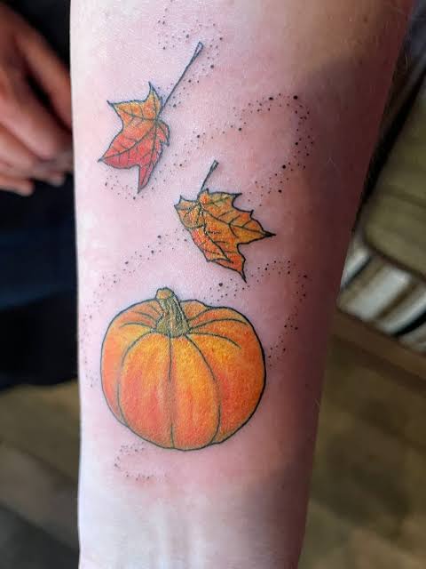 A cheerful orange pumpkin sits below two falling leaves in a colorful forearm tattoo.