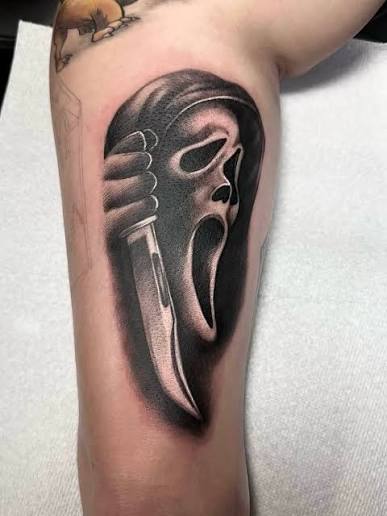 Black shaded upper arm tattoo of Ghostface with an open mouth beside a long knife.