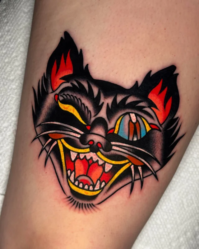 Thigh tattoo of a grinning black cat with one eye shut and the other filled with color.