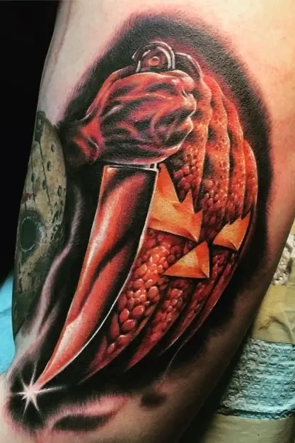 Upper arm tattoo of a gleaming knife beside a grinning pumpkin, with red shading and black shadows.