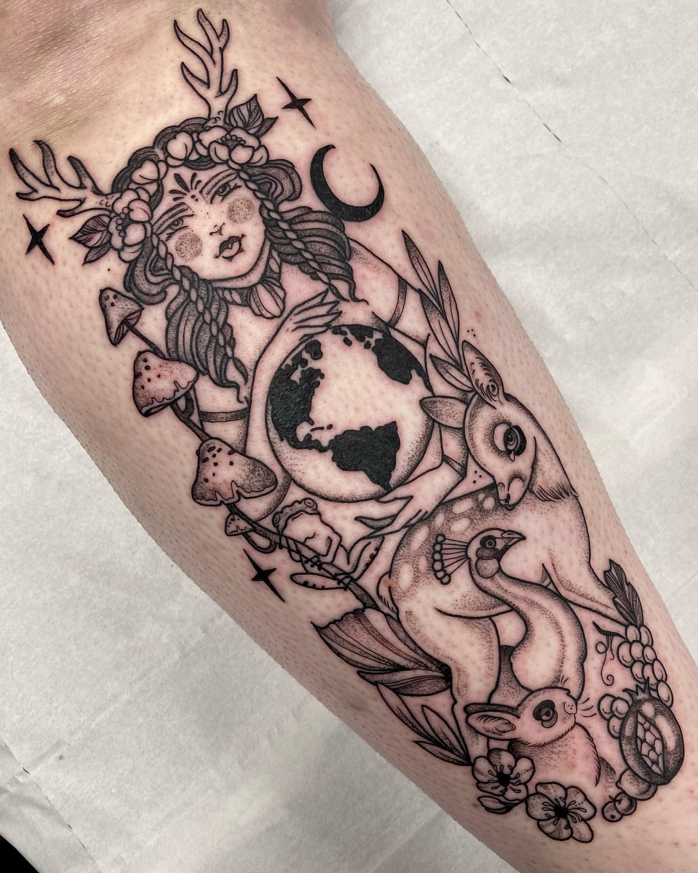 Antlered woman holding the Earth beside a deer, rabbit, and mushrooms in a shaded leg tattoo