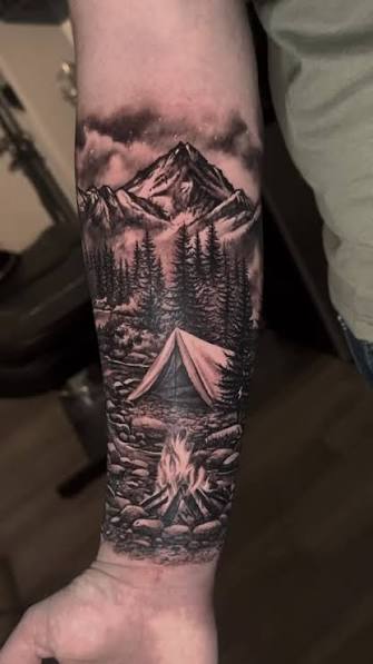 Black and gray forearm tattoo of a campfire and tent among pines below mountains