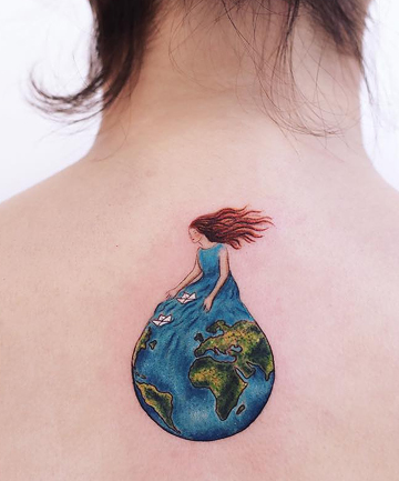 Windswept woman in a blue dress sitting on the globe in an upper back tattoo