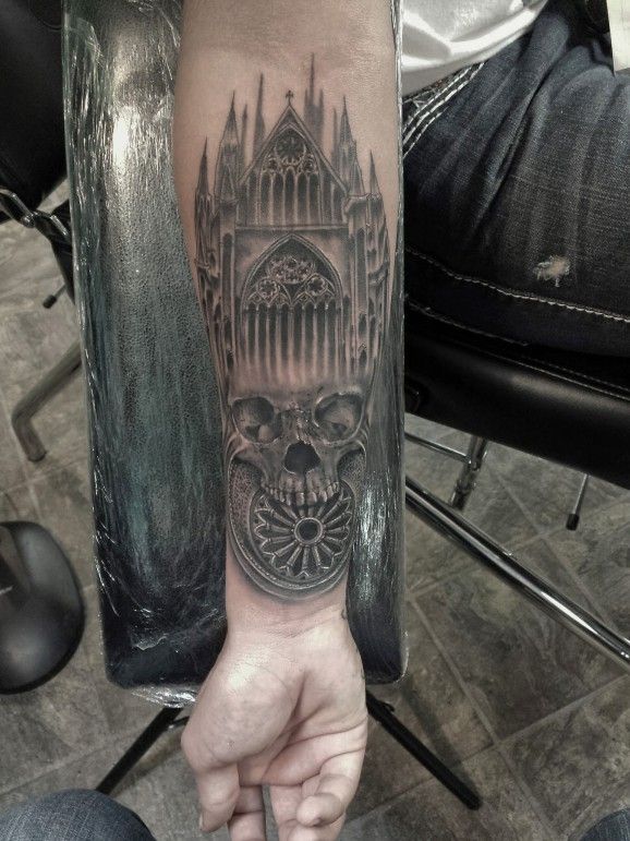 Skull, rose window, and cathedral spires arranged in a black and gray inner forearm tattoo
