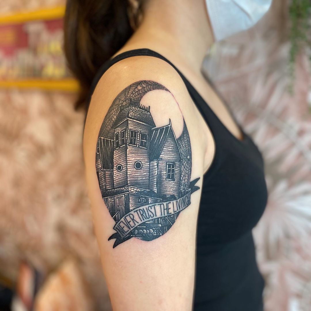 Banner script haunted house tattoo on the upper arm shows a crooked house in a web lined oval with a warning ribbon below.