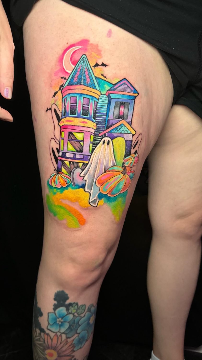 Rainbow watercolor haunted house tattoo on the thigh shows a sheet ghost drifting past a pastel Victorian house.