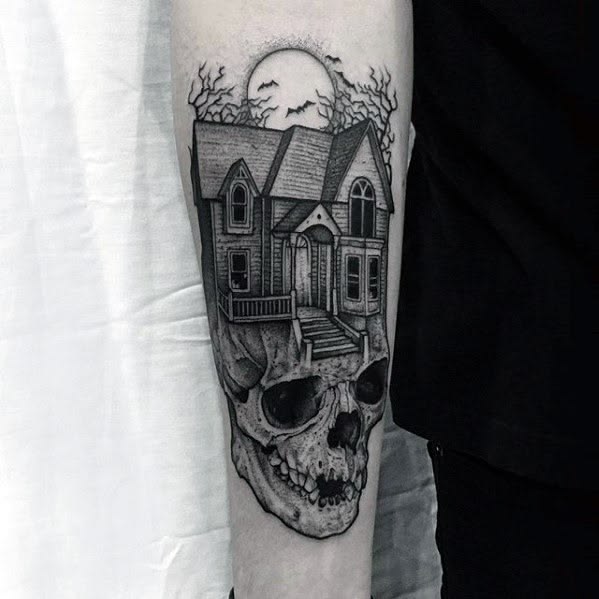 Skull and haunted house forearm tattoo shows a crooked mansion rising from a cracked skull under a full moon with bats.