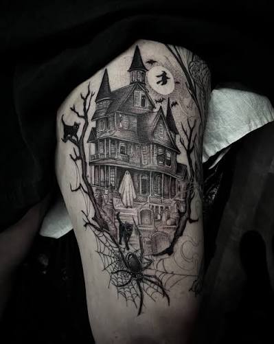 Bold black thigh tattoo shows a crooked Victorian mansion, bare trees, and a witch flying past a full moon.