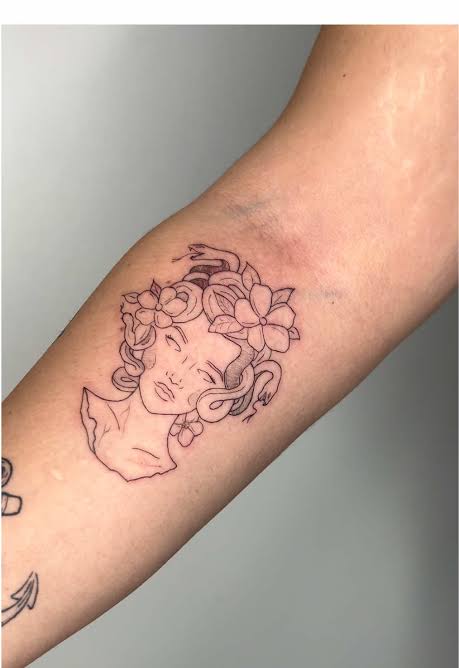 Fine line inner arm tattoo depicting Medusa with closed eyes, flowers, a snake crown, and minimal shading.