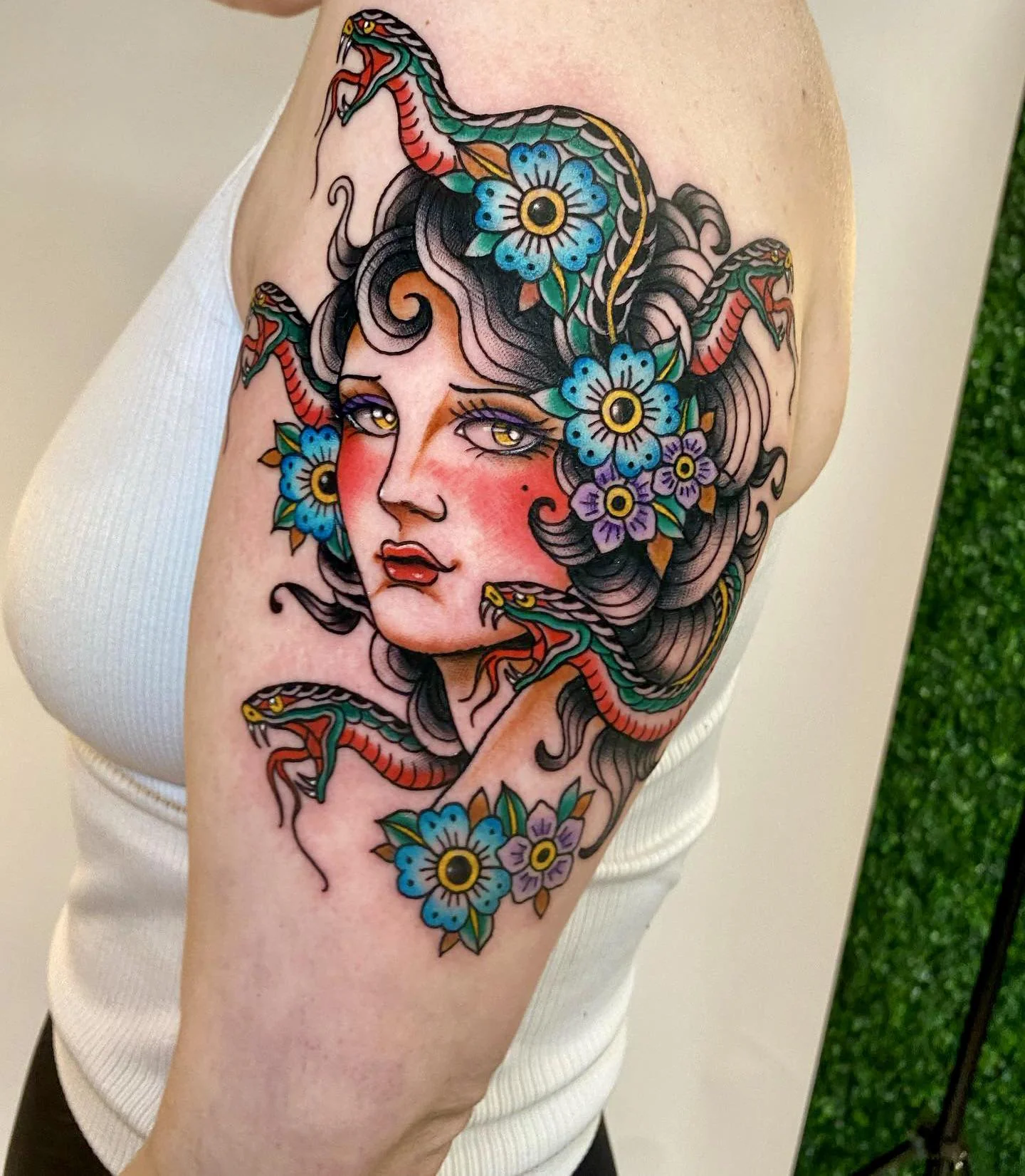 Traditional style Medusa upper arm tattoo with blue flowers, green snakes, wavy hair, and bold outlines.