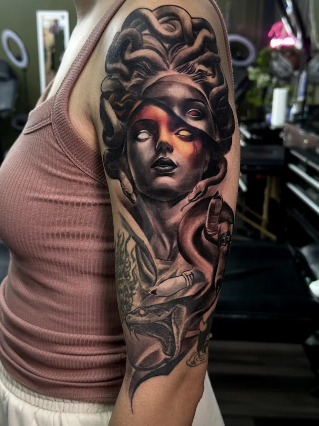 Upper arm tattoo of Medusa with a tilted mask, fiery light across her face, thick snake coils, and an open jaw below.