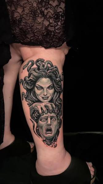 Darkly shaded thigh tattoo showing a smiling Medusa holding a screaming severed head beneath her face.