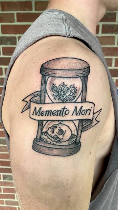 Outer upper arm tattoo of an hourglass containing a tree above a skull, wrapped with a memento mori banner.