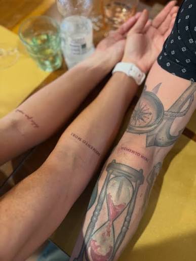 Matching small memento mori script tattoos on arms, with an hourglass tattoo nearby on one arm.