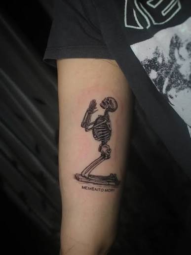 Kneeling skeleton tattoo with hands held in prayer and tiny memento mori lettering beneath its knees on the upper arm.