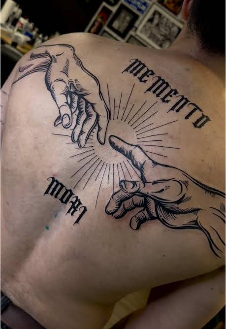Upper back tattoo with large reaching hands, rays around the fingertips, and memento mori in Gothic script.