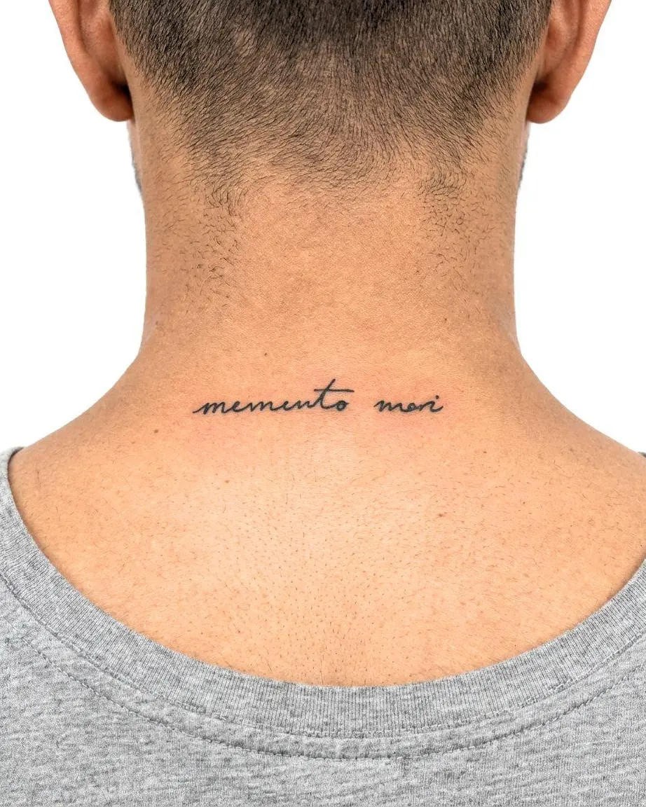 Small memento mori tattoo in flowing black script across the base of the neck above the shirt collar.