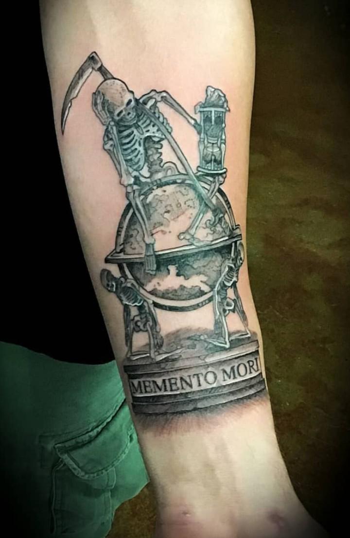 Black and grey forearm tattoo of a skeleton seated on a globe with an ornate stand, beside a scythe and hourglass.