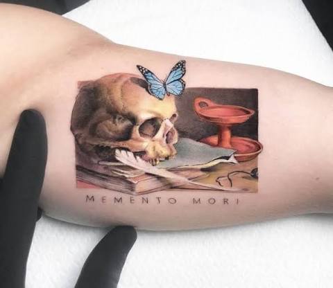 Colorful rectangular inner upper arm tattoo showing a blue butterfly perched on a skull in a dark still life.