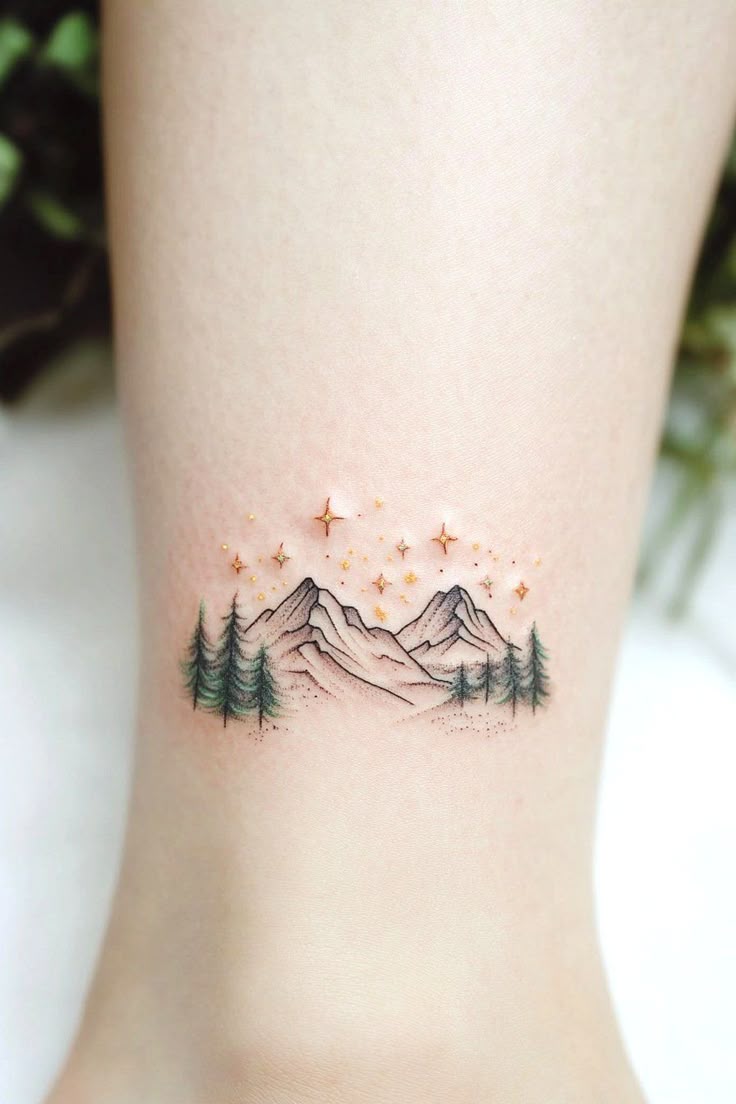 Slim mountain and pine tree tattoo with tiny gold stars above the ankle