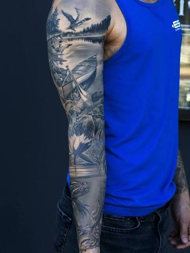 Birds fly over a lake and pine trees above large flowers on a black and gray tattoo sleeve.