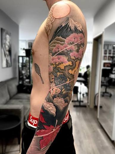 Red maple leaves and pink blossoms surround a dark mountain scene on a tattoo sleeve.