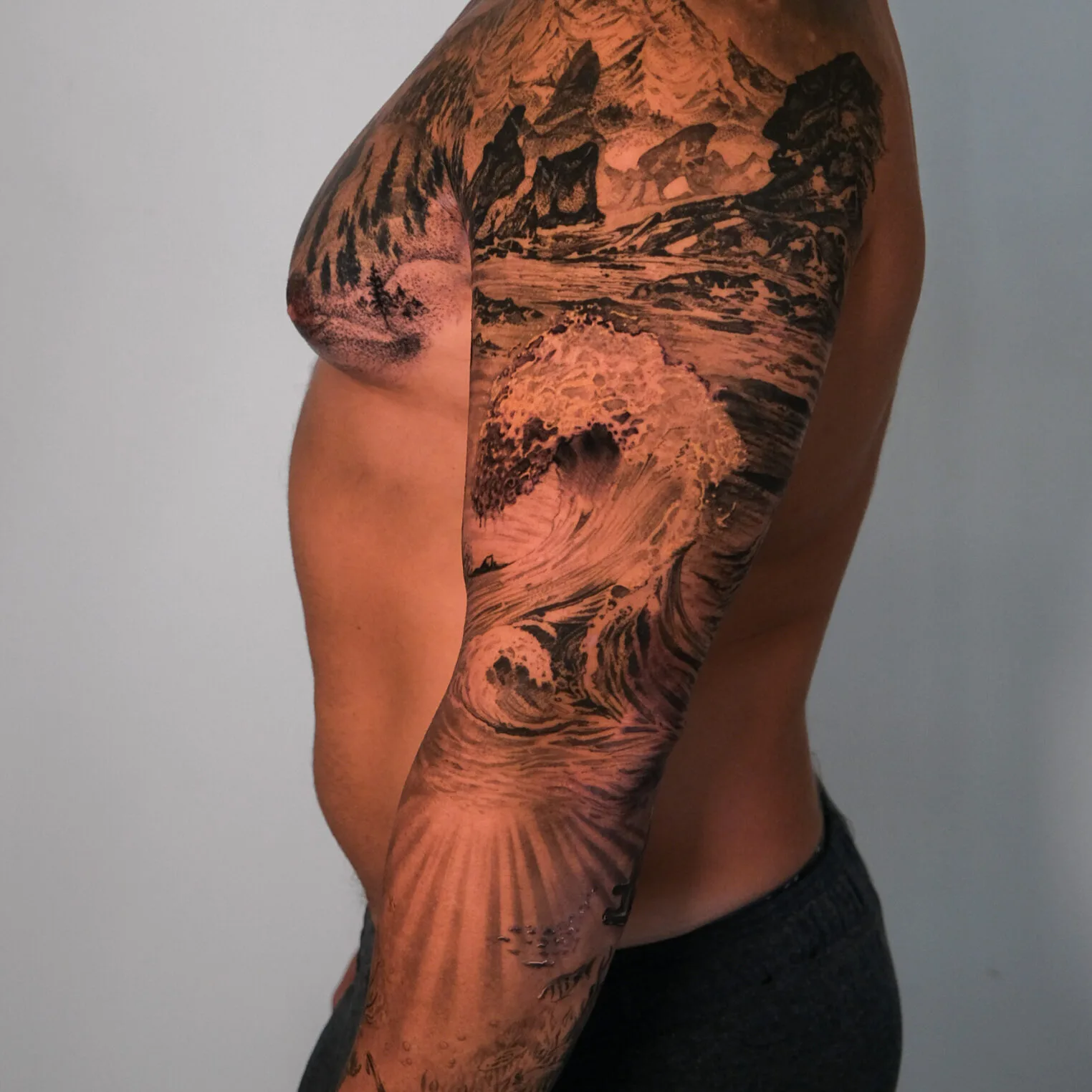 Rocky mountains rise above a crashing ocean wave on a dark tattoo sleeve.
