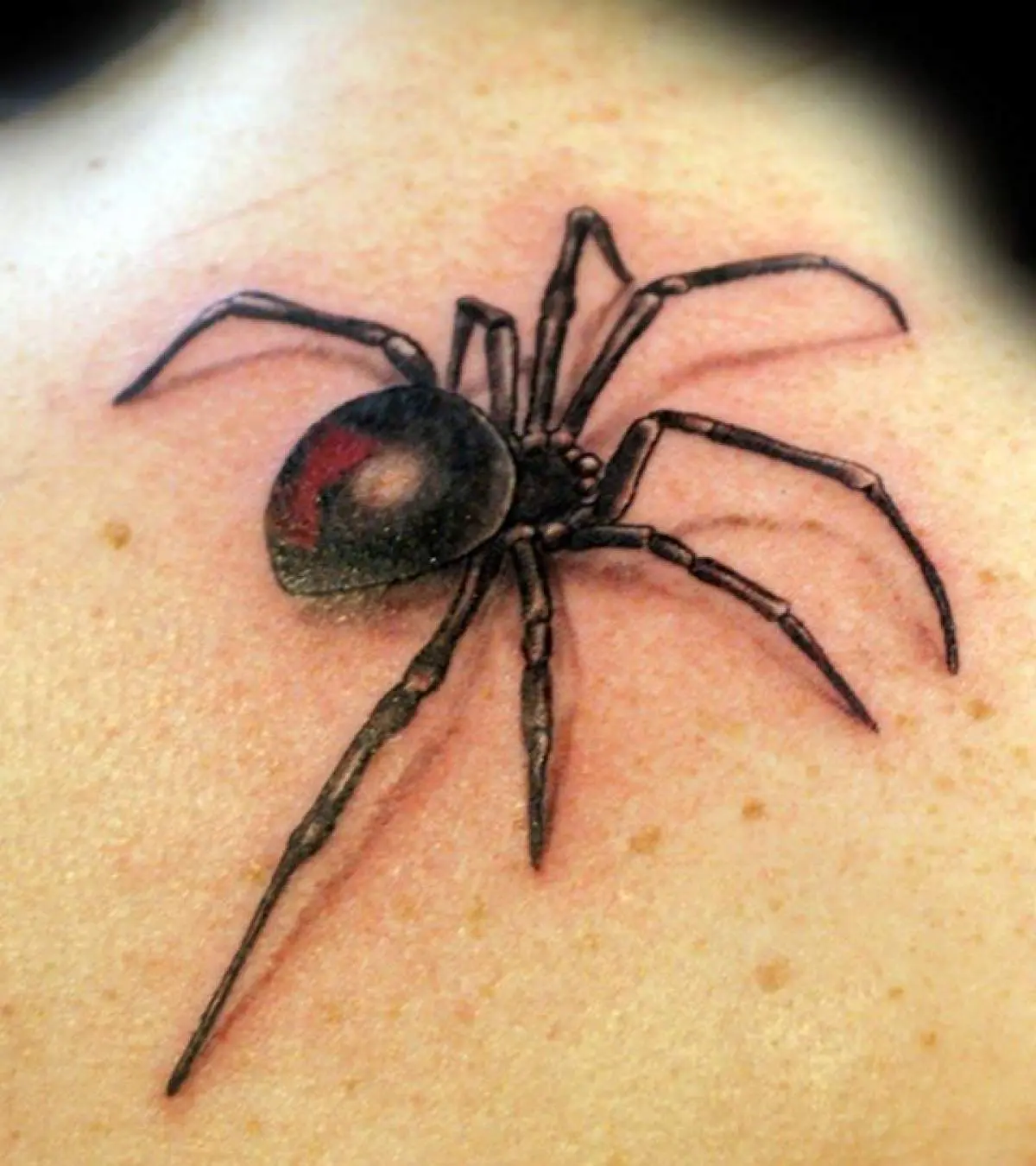 Realistic black widow spider tattoo with a red hourglass near the collarbone and shoulder.