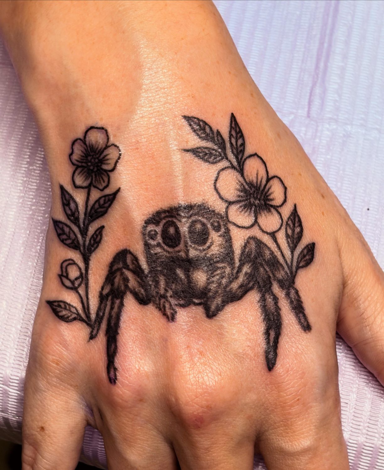 Cartoon jumping spider with big eyes tucked between two flowers on the hand.