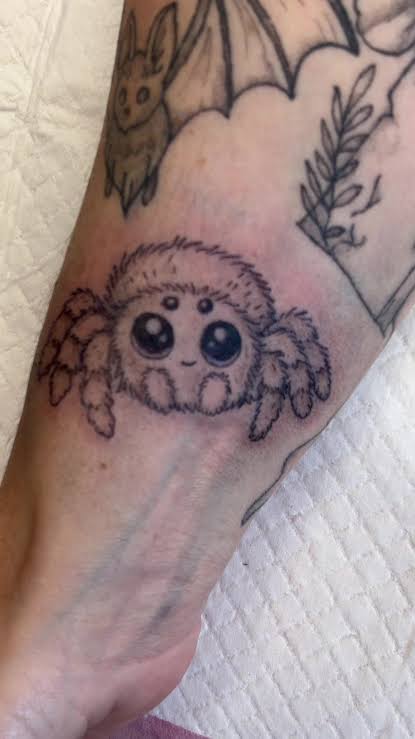 Fuzzy baby spider with glossy eyes near a bat outline on the wrist.