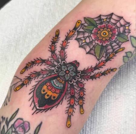 Neo traditional spider tattoo in teal and orange with a flower centered web on the inner elbow.