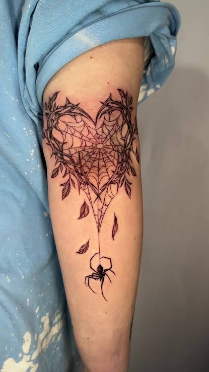 Heart shaped spiderweb tattoo with a small spider hanging from thread, wrapped in leafy vines on the upper arm.