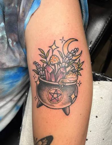A tiny witch tattoo cauldron filled with crystals, mushrooms, and a frog under a crescent moon on the upper arm.