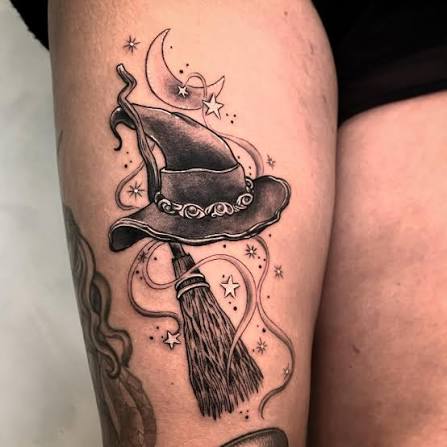 A witch hat and broom floating above pink swirls, a crescent moon, and tiny stars on the thigh.