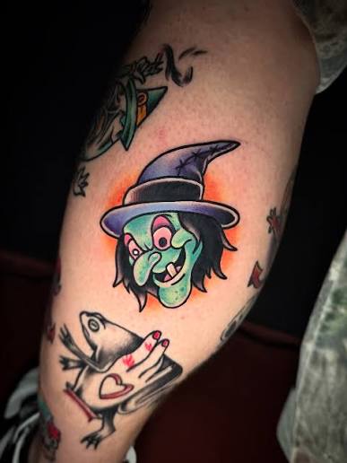 A green faced witch tattoo with red eyes and a purple hat next to a frog holding a spellbook on the calf.