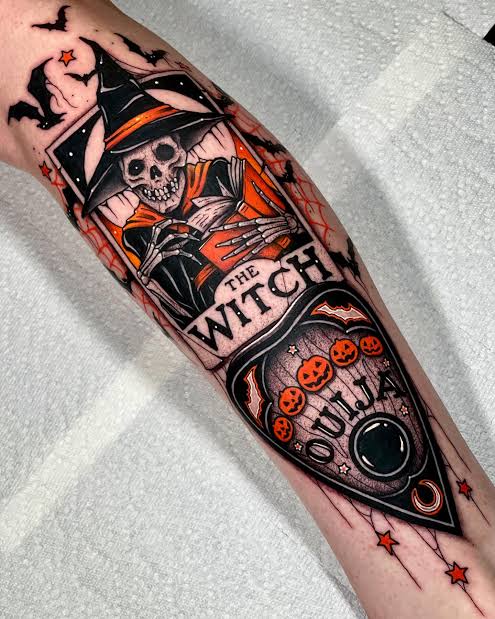 An orange and black skeleton witch tattoo card with bats, spiderwebs, and a glowing planchette on the forearm.