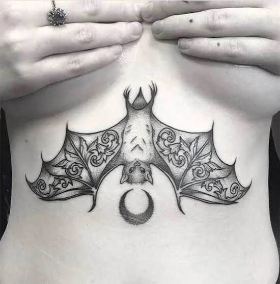 A lacy bat tattoo with a crescent moon in the underboob spot, shaded in soft gothic tones.