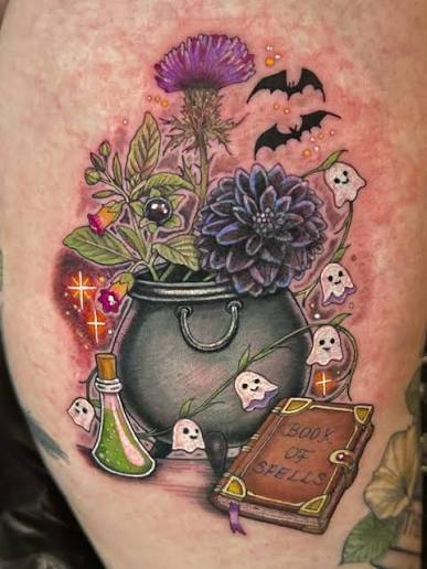 A witchy garden tattoo with a cauldron, thistle, dahlias, ghost faces, bats, and a spellbook on the thigh.