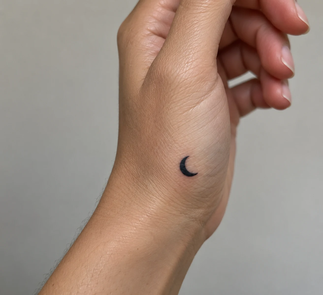 Tiny solid black crescent moon tattoo near the outer edge of a hand.