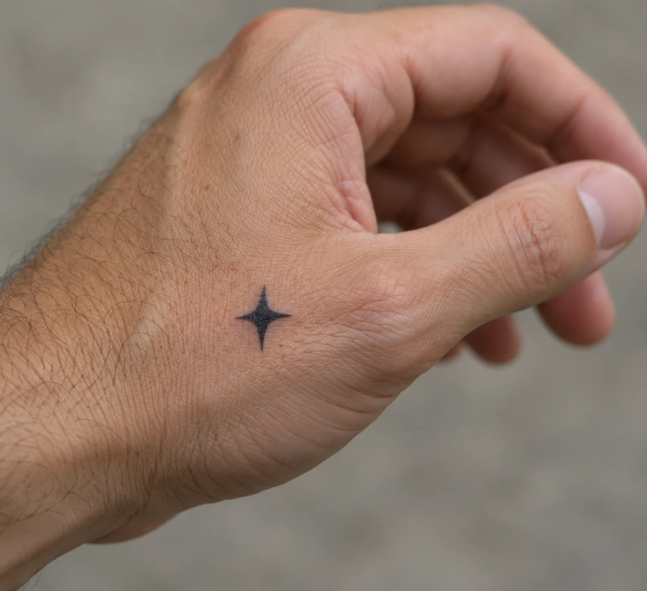 Small four-point black star tattoo near the thumb base on a man's hand.