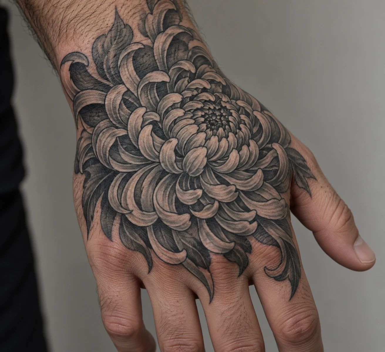 Black and grey chrysanthemum tattoo with layered curved petals covering the back of a man's hand.