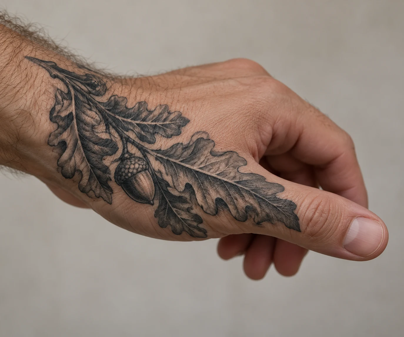 Black and grey oak leaves with one acorn tattooed across the thumb side of a man's hand.
