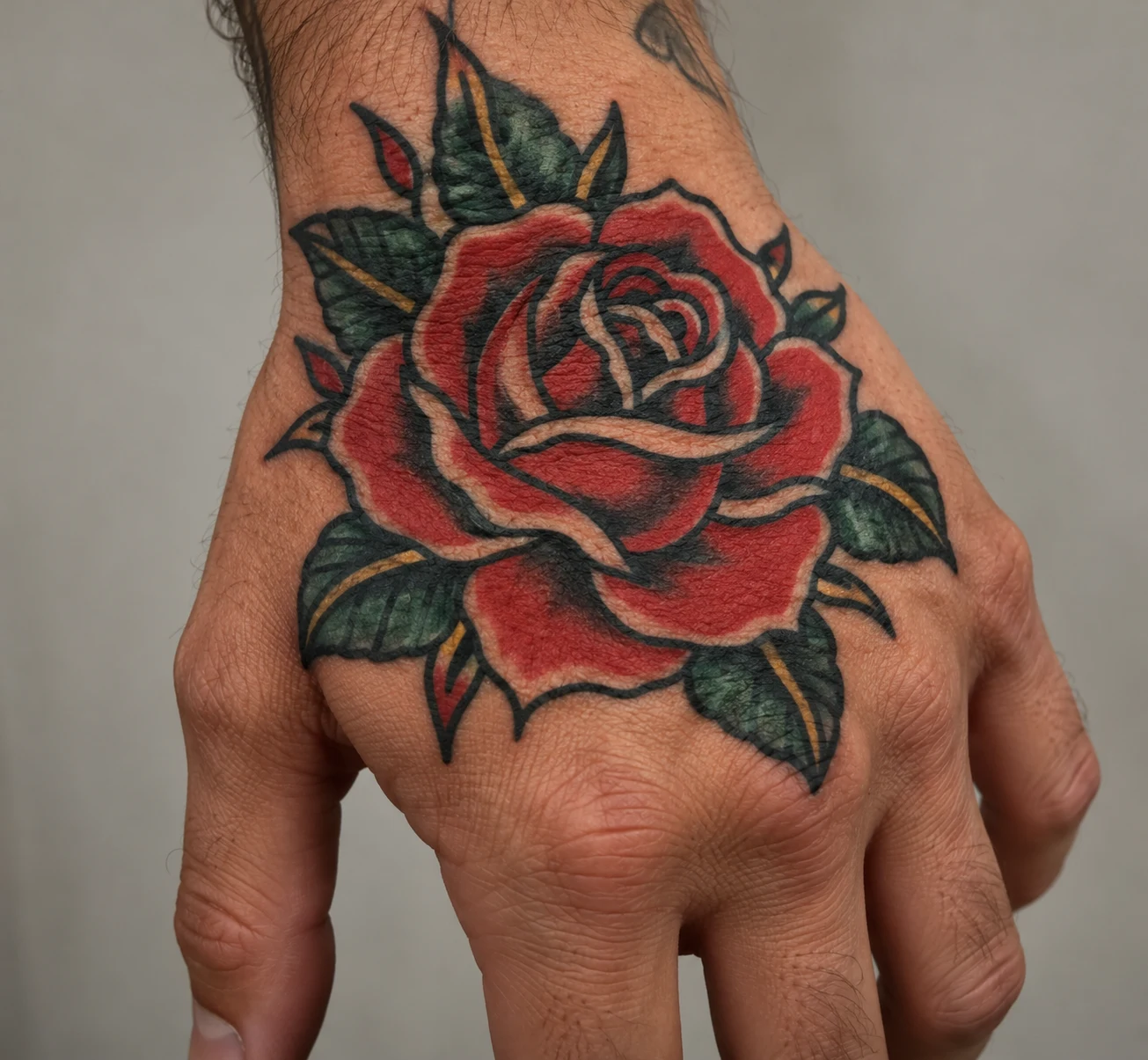 Traditional red rose tattoo with bold black outlines and green leaves on the back of a hand.